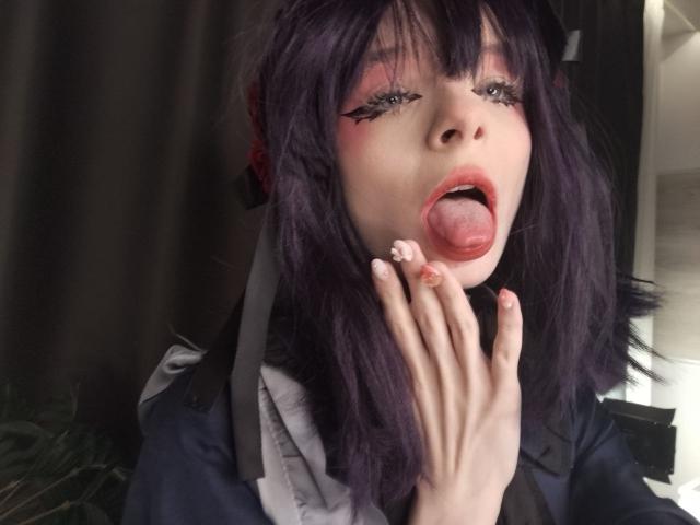 ChangCaddle has purple hair, pink eyeshadow, and is sticking out her tongue with her hand near her mouth. Her nails are decorated. She's wearing a dark jacket.