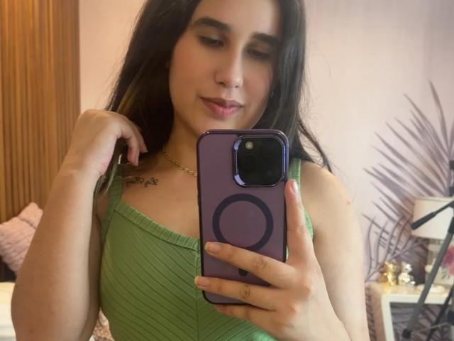 KatieDesirX usando uma blusa verde canelada, segurando um telefone roxo, mostrando seios pequenos, cabelo preto longo, piercing no nariz, sorriso discreto, mão perto do queixo, colar de ouro, pequena tatuagem no peito esquerdo..