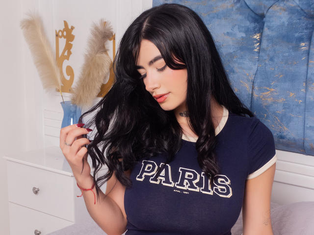 Bethani Williams mit schwarzen, welligen Haaren, einem blauen "PARIS"-Shirt und rotem Lippenstift, berührt sanft ihre Haare und zeigt dabei ihre großen, runden, festen Brüste. Sie befindet sich in einem weißen Raum mit blauen Kunstwerken..
