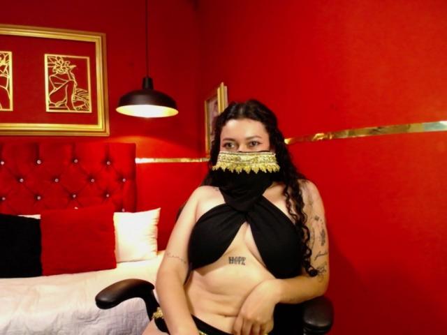 AlanaNaur with long dark curly hair, wearing a black mask and draped top, exposing her big tits and belly tattoo, sitting in a red room.