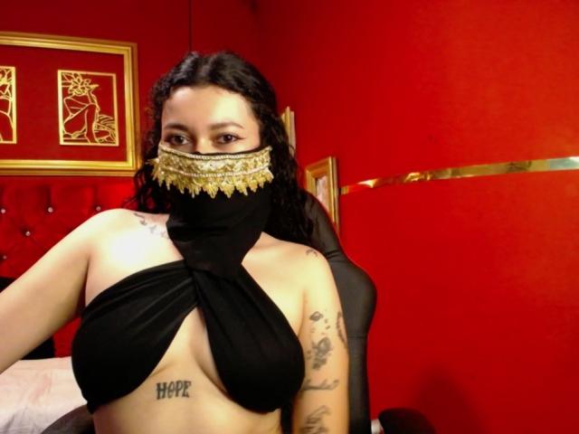 AlanaNaur with dark curly hair, wearing a black mask with gold trim, black halter top exposing her tits and "HOPE" tattoo, in a red room.