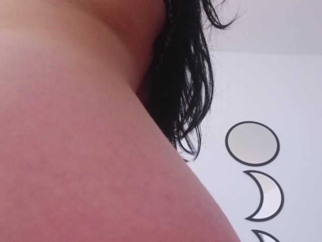 ZarayFeet's side boob with a small, dark tattoo of a crescent moon and circle on a white wall, showing black hair partially in frame.