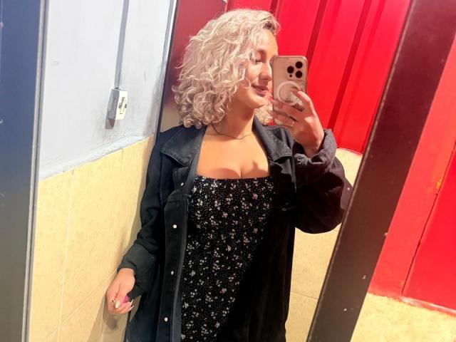 ElenaSaenz with curly gray hair, black floral dress, and black coat, taking a mirror selfie in a red-walled room, showing her tits slightly under the dress.