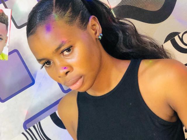 Nastybitch-hot with dark skin, braided hair, black tank top, small earrings, purple and green face paint, serious expression, visible ass, medium-sized tits, and a reflection in the upper left.
