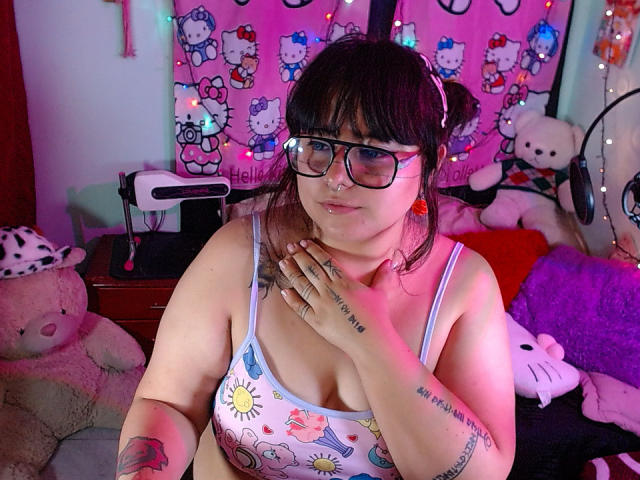 LilithHeavy with glasses, nose ring, and tattoos, wearing a sun and moon print tank top, showing her tits, hand on chest, in a colorful room with stuffed animals.