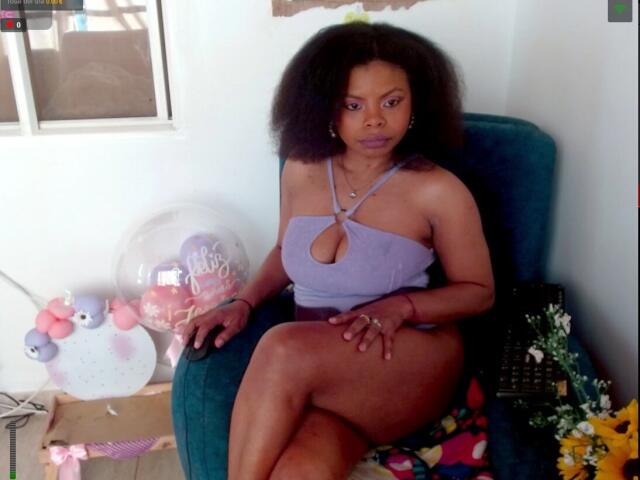LizReyS with dark brown curly hair, wearing a lilac halter top with a cleavage cutout, sitting in a blue chair, showing her ass, with a bouquet on the right.
