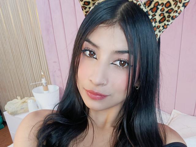 NaiaSanders with black hair, brown eyes, and pink lips, wearing a leopard print headband, nose ring, and earrings, smiling at the camera, showing her bare shoulders.