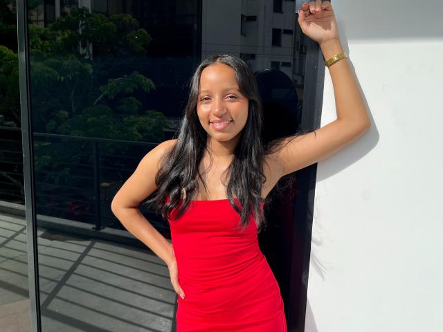 RaquelNoir with long black hair, tan skin, and a cute smile, wearing a tight red dress, standing against a white wall, hand on hip, other arm raised.