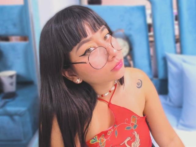 Genesiss with long black hair, round glasses, red top showing her tits, and a small tattoo on her right shoulder, pouting with pink lipstick, sitting in a blue room.
