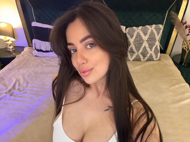 ArielGemme with long brown hair, blue eyes, and pink lips, wearing a white top, showing her tits, sitting on a bed with a green headboard.