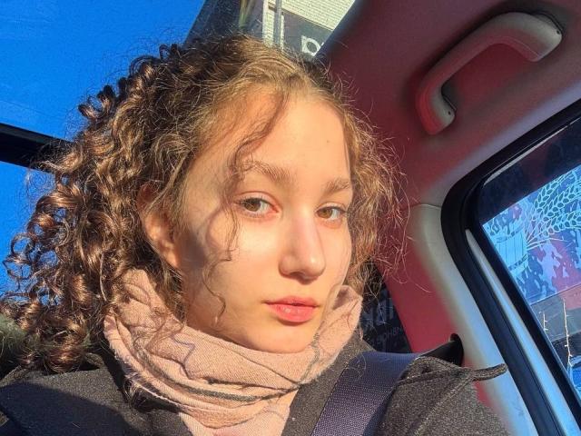 ShantaeTsuha with curly brown hair, light skin, and green eyes, wearing a pink scarf and black jacket, seated in a car, sunlight on her face, looking at the camera.