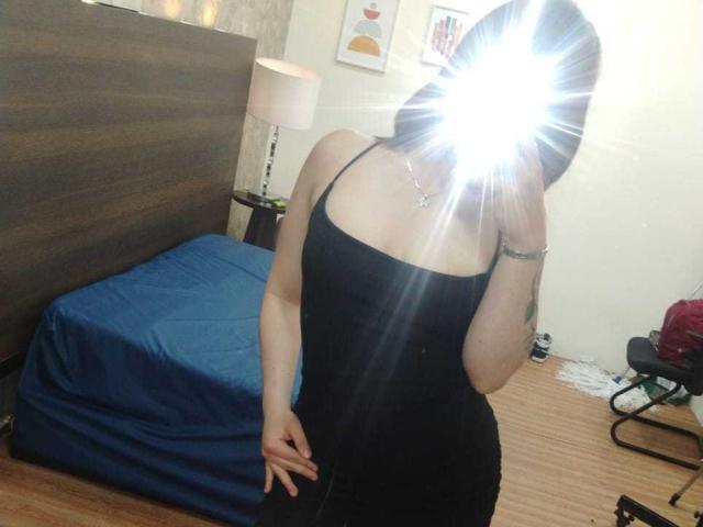 Mia Colemann, vestindo um vestido preto com alças finas, com o flash da câmera sobre o rosto, exibindo suas nádegas, seios pequenos e uma tatuagem no braço direito, está em pé em um quarto..