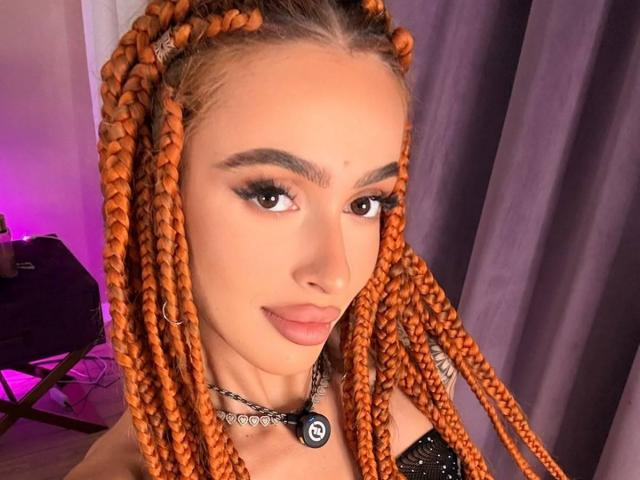 MarissaFex with orange braided hair, full lips, and thick eyebrows, wearing a black choker with a blue emblem, has a cute, sexy look.