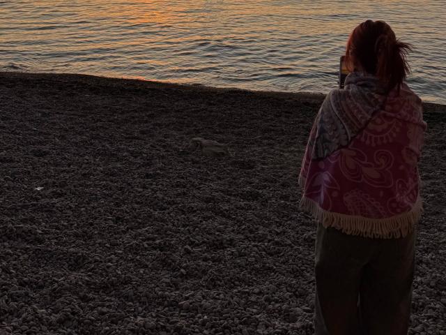 AlexXas stands on a pebbly beach with a red and pink patterned shawl, facing a calm water surface at sunset. She's wearing green pants and has her back to the camera.