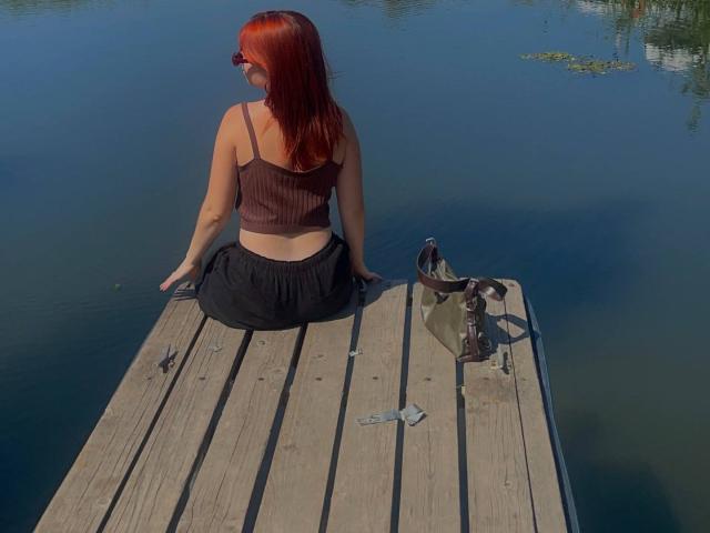 AlexXas with red hair sits on a wooden dock, facing water, wearing a brown crop top and black skirt, with a green bag and sunglasses beside her.