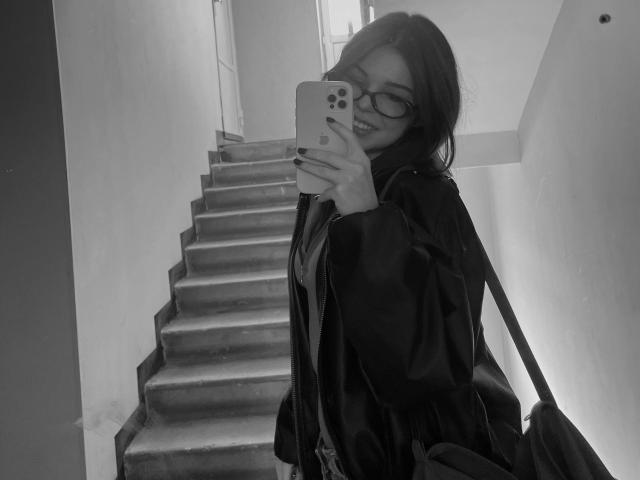Black-and-white selfie of AlexXas with glasses, dark hair, and a shiny black jacket, holding a phone with a colorful case, standing on a staircase, showing her ass.