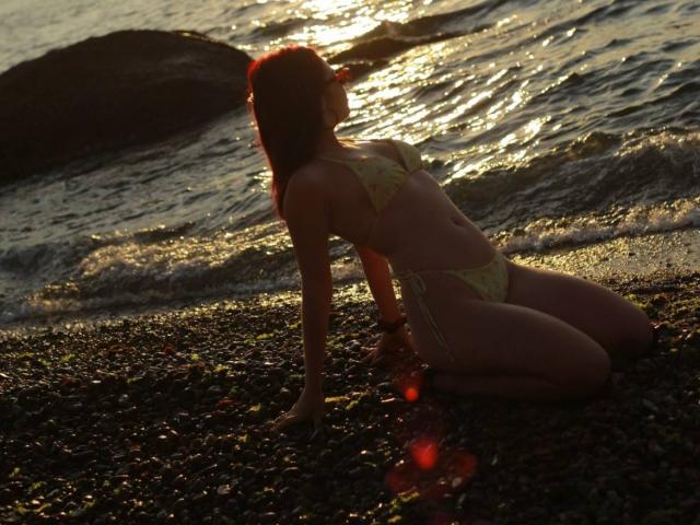 AlexXas in a yellow bikini, kneeling on a rocky beach, back to the camera, sun setting, water reflecting, lens flare, small tits, slim, brown hair, wet skin.