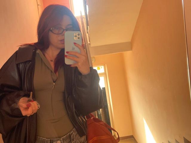 AlexXas with red hair, glasses, green top, black leather jacket, holding phone, mirror selfie, showing small tits, slightly visible ass, holding phone in right hand, door in background.