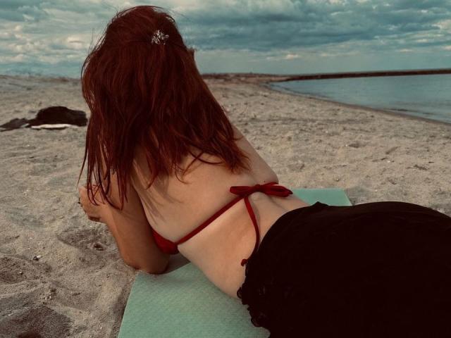 AlexXas is lying on a green mat, facing away, in a red bikini top and black bottoms, with red hair, on a sandy beach, under a cloudy sky.
