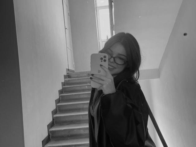 Black-and-white selfie of AlexXas with glasses, smiling, holding phone, wearing dark jacket, standing on stairs, showing tits slightly visible under jacket.