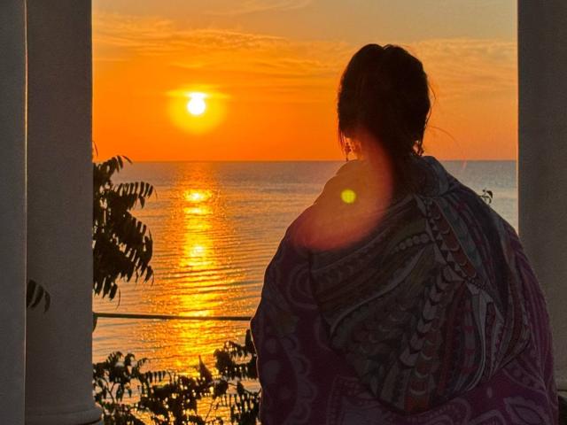 AlexXas with dark hair in a patterned purple and blue shawl, sitting with back to camera, watching a bright orange sunset over a reflective ocean.