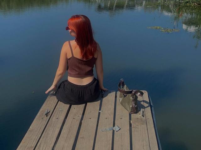 AlexXas with red hair sits on a wooden dock, facing a calm water body, wearing a brown tank top and black skirt, with a brown bag to her right.