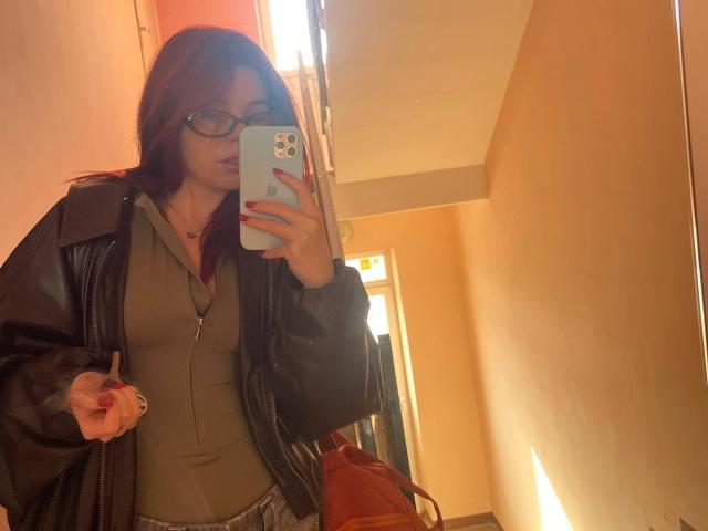 AlexXas with red hair, glasses, and black leather jacket, holding phone, showing tits in green top, red nails, and light skin, standing in sunlit room, mirror selfie.