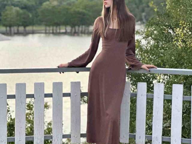 Eugenia Vanbecelaere, com cabelos castanhos longos, vestindo um vestido marrom de manga comprida, está em pé em uma varanda de madeira branca, com as mãos na grade. Ao fundo, há árvores verdes e água..