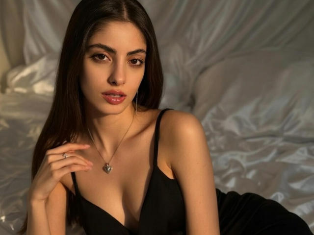 EugeniaVanbecelaere has long dark hair, brown eyes, and is wearing a black dress with a low neckline, showing her small tits. She's on a bed with white sheets.