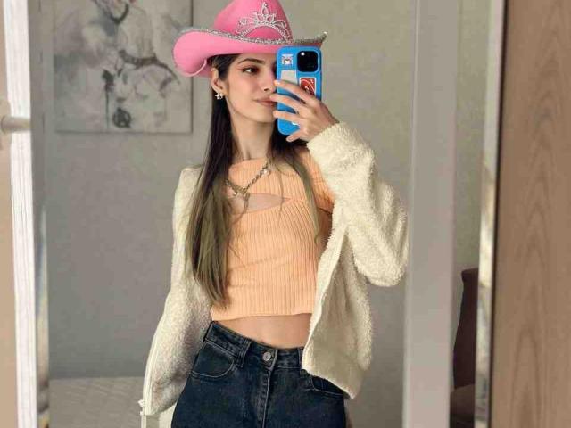 EugeniaVanbecelaere with pink cowboy hat, white fuzzy cardigan, orange ribbed crop top, and dark jeans, holding blue phone, showing slim waist and long green-tipped brown hair.