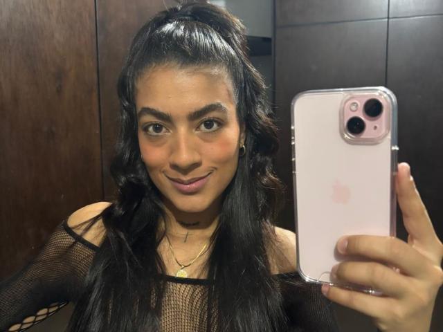 ZanderAndZanotti, avec de longs cheveux noirs attachés en queue de cheval, souriant, portant un haut en filet noir, tenant un iPhone rose, prenant un selfie devant un miroir, montrant un visage mignon et des seins..