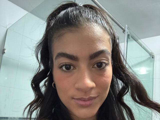 ZanderAndZanotti has dark brown eyes, thick eyebrows, long wavy black hair in a ponytail, and wears small gold earrings with a slight smile, showing a cute face.
