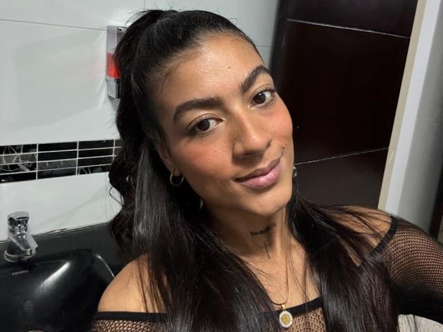 ZanderAndZanotti tiene el cabello oscuro recogido en una coleta, ojos marrones y una ligera sonrisa. Lleva una blusa de malla negra que deja ver sus pequeños senos, y tiene un tatuaje debajo del mentón..