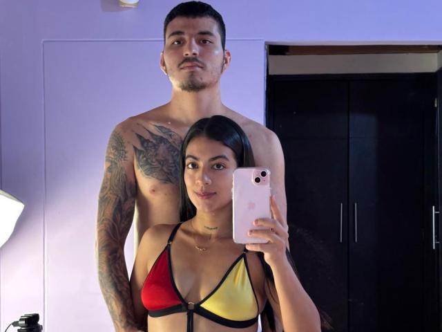 ZanderAndZanotti with small tits and a red-yellow-black bikini stands in front of ZanderAndZanotti, shirtless with tattoos, holding a phone. ZanderAndZanotti's arm is around ZanderAndZanotti's waist.