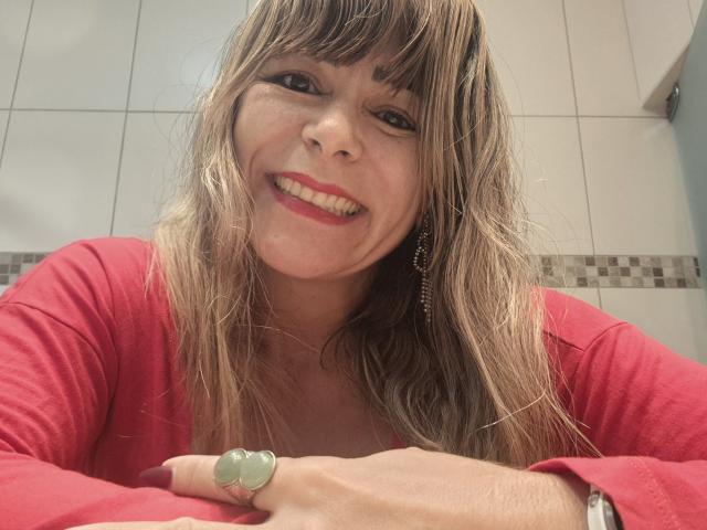 CarollineBer with wavy blonde hair, red lipstick, and a green ring, smiling at the camera in a pink top, wearing a chain earring, and a watch.