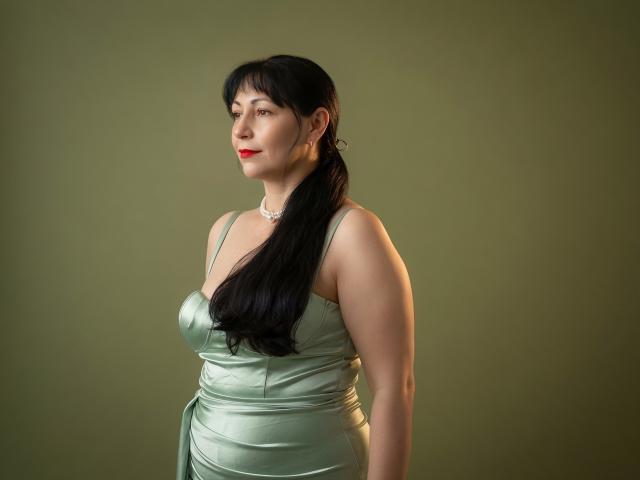 LiluGrants with long black hair in a ponytail, wearing a shiny green satin dress, red lipstick, and a pearl necklace, standing against a green background. Boobs visible.
