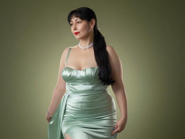 LiluGrants has large tits, wears a shiny green satin dress, long black ponytail, red lipstick, silver necklace, and green background. She's looking to the side with a slight smile.