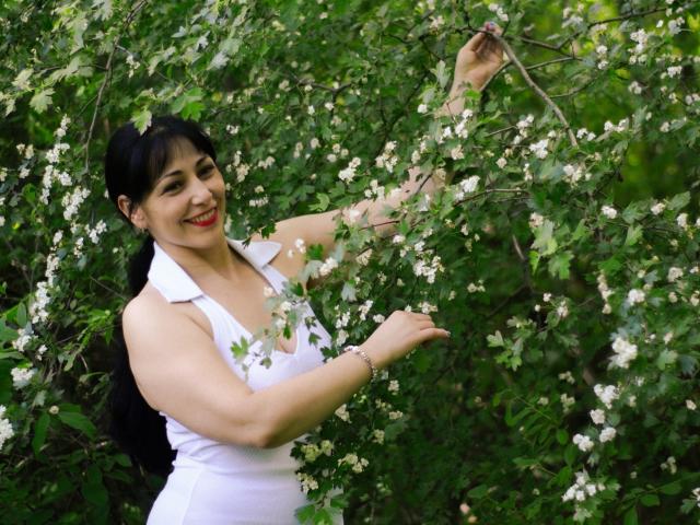 LiluGrants with black hair, white sleeveless top, and red lipstick, holding a branch with white flowers, smiling, showing her tits, surrounded by green foliage and flowers.