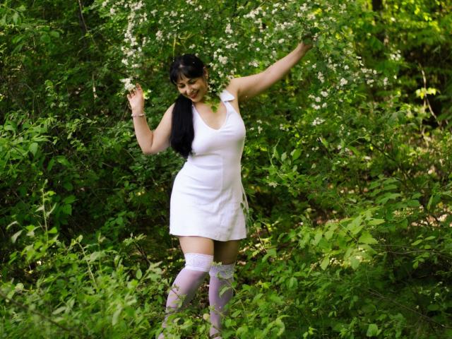 LiluGrants mit langem, schwarzem Haar, einem weißen Kleid und rosa, hohen Strümpfen, steht in einem grünen Wald, die Arme erhoben, lächelnd und nach unten blickend, umgeben von weißen Blumen und Grün..