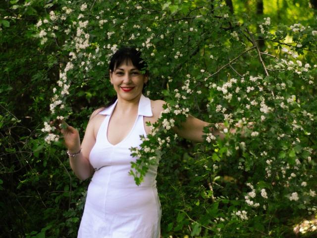 LiluGrants has curly black hair, red lipstick, and a white sleeveless dress, standing amidst green foliage and white flowers, smiling with arms slightly raised. Her tits are visible.