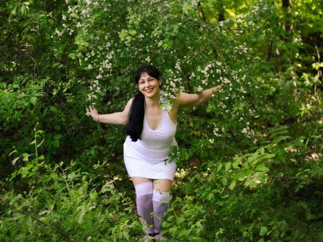 LiluGrants with black hair, white dress, and white thigh-highs, smiling with arms out, standing in green foliage, showing her big ass and large tits.