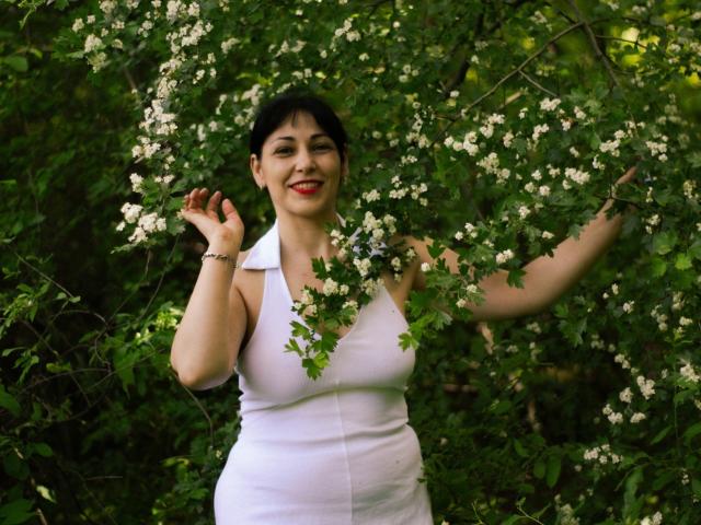 LiluGrants with dark hair, red lipstick, and white sleeveless dress, holding white flowers, smiling, arms raised, standing in green foliage. Boobs visible, curvy, medium-sized.