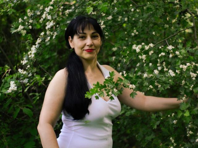 LiluGrants with black pigtails, red lipstick, white tank top, and medium tits, standing among green leaves and white flowers, smiling softly at the camera.