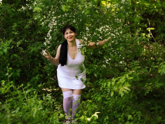 LiluGrants has big tits, wearing a white dress and purple thigh-highs, standing in greenery with white flowers, smiling with black pigtails, arms raised.