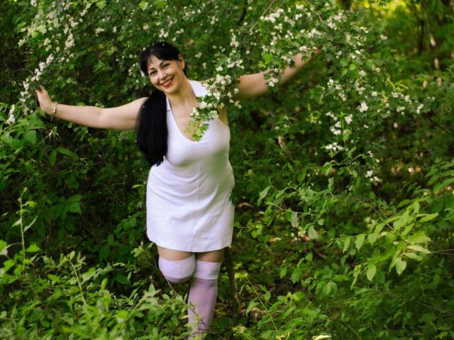 LiluGrants with black hair, smiling, arms out, wearing white dress and pink thigh-highs, standing in green foliage, surrounded by white flowers, showing her big tits.