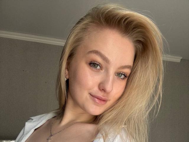 NievesMegee has blonde hair, green eyes, and a cute smile, wearing a white top, with a silver necklace, and small earrings, in a well-lit, soft-focused selfie.