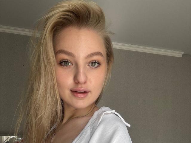 NievesMegee with blonde hair, green eyes, and a slight smile, wearing a white shirt, looking at the camera with a soft, natural light. Her tits are partially visible.