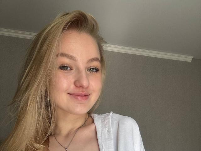 NievesMegee a les cheveux blonds, les yeux verts et un joli sourire avec des lèvres roses. Elle porte un haut blanc et un collier en argent, et regarde légèrement vers la droite..