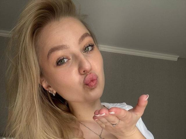 NievesMegee, avec ses yeux verts gonflés, ses cheveux blonds et ses lèvres roses, faisant une moue de baiser, tenant sa main sans alliance, avec des ongles blancs, portant un haut blanc, dans un autoportrait..
