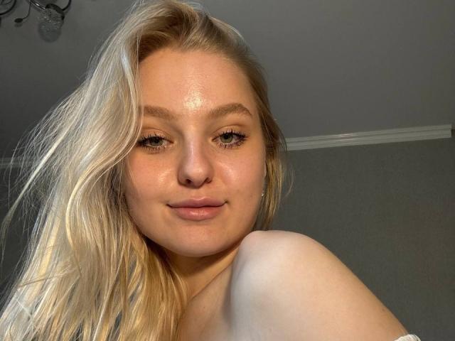 NievesMegee has shiny, blonde hair, light brown eyes, and a subtle smile, with her bare shoulder and a slightly glossy face visible in the image.